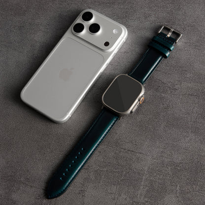 A dark teal leather watch strap with silver hardware, laid next to a silver smartphone on a textured gray surface.