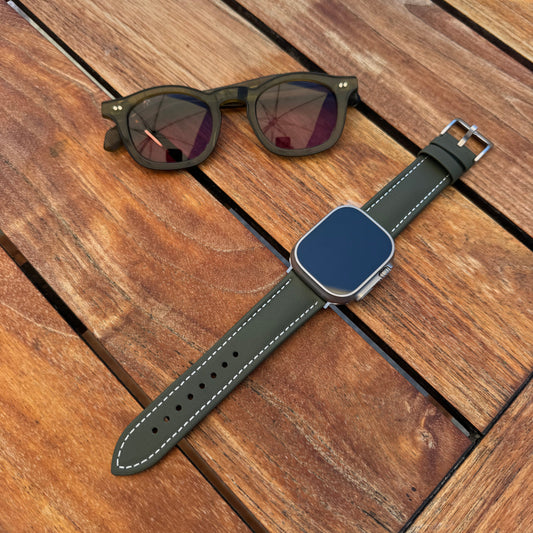 The Helm Olive Green Sailcloth Apple Watch Strap resting on a wooden table beside sunglasses