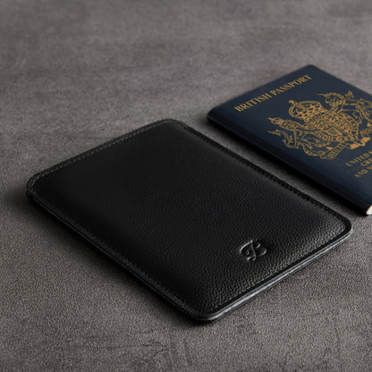 Black leather passport sleeve beside a British passport on grey surface – The Bridge