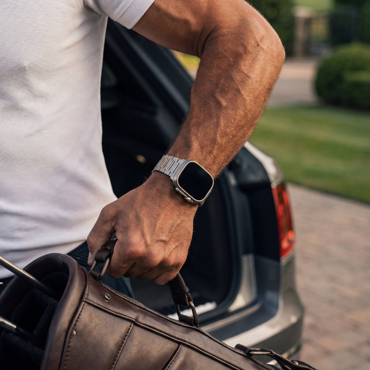 The Presidential titanium Apple Watch strap on a wrist, lifting a leather holdall from a car
