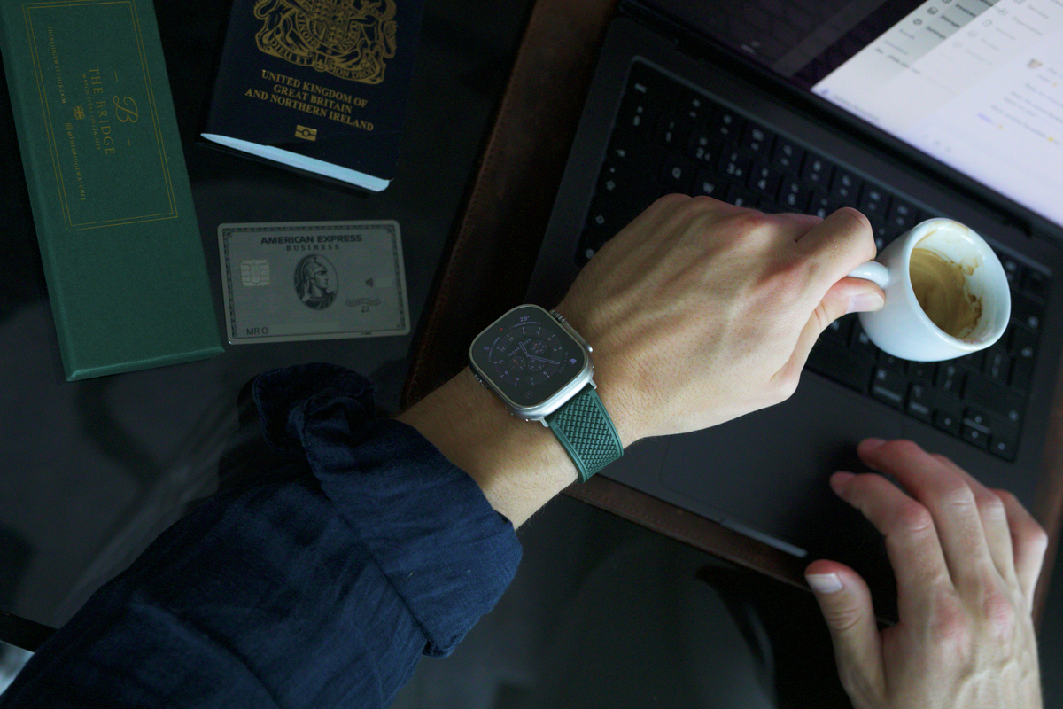The Aurelius Apple Watch strap on a wrist at a desk with laptop, coffee and passport