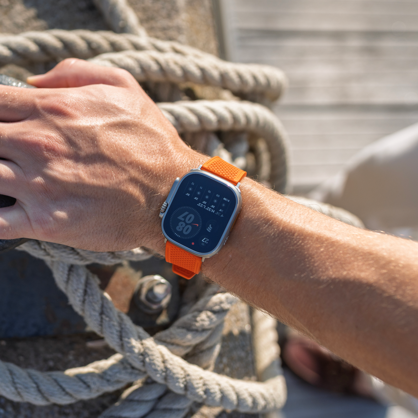 Person wearing a smartwatch with an orange band, interacting with ropes on a dock.