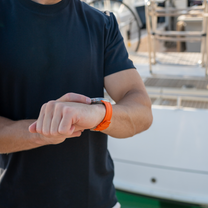 Person wearing a black shirt and white pants, checking their orange watch with a boat in the background.
