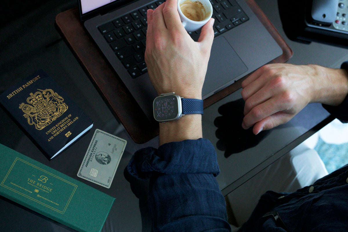 The Aurelius Apple Watch strap in navy on a wrist at a desk with laptop, coffee and passport